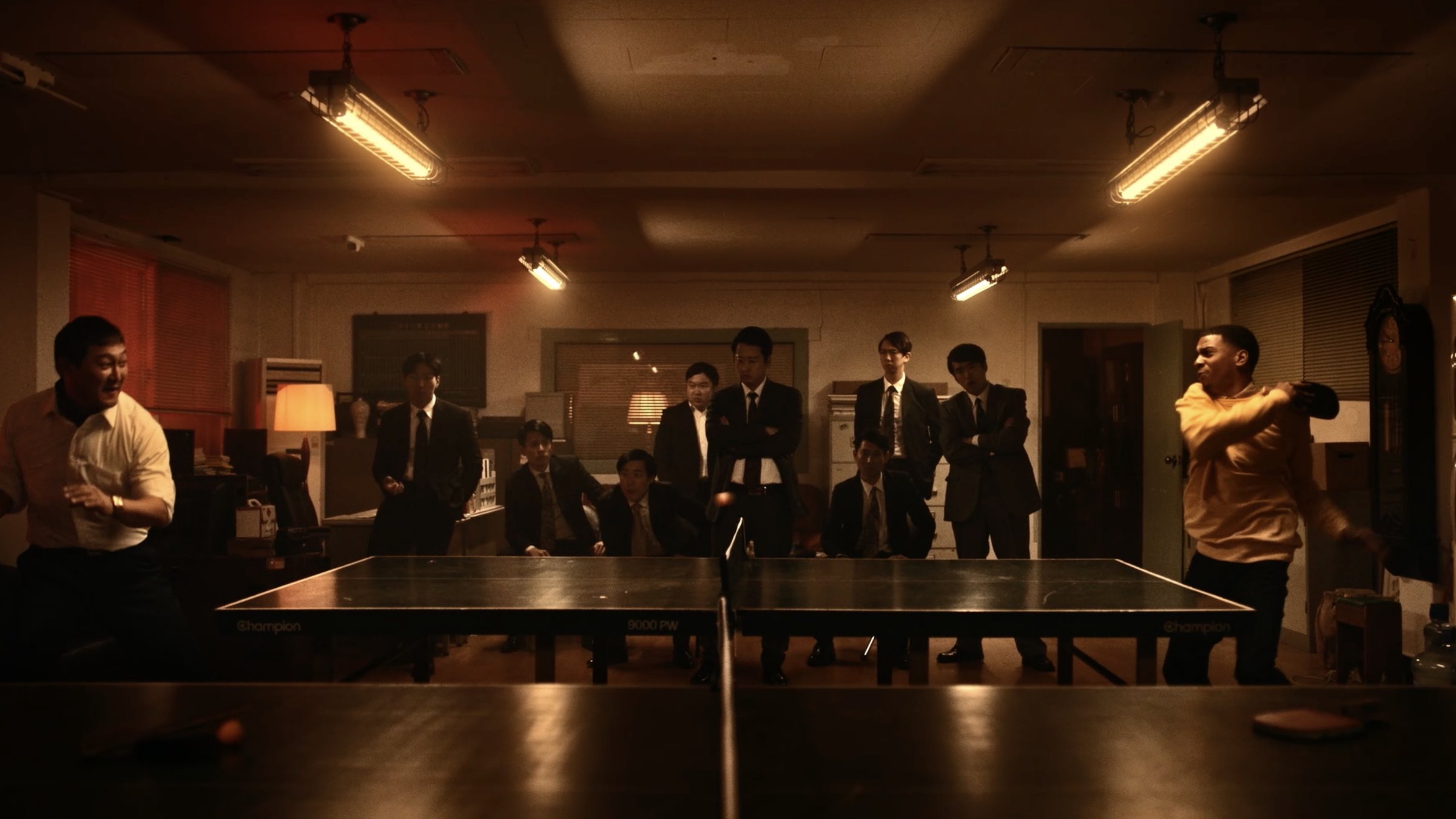 A ping-pong showdown in a smoky office, watched by a row of men in dark suits