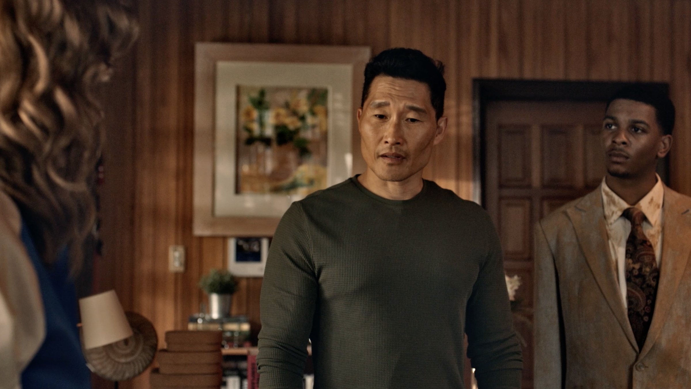 A man in a green sweater and a man in a dust-covered suit stand in a wood-paneled room