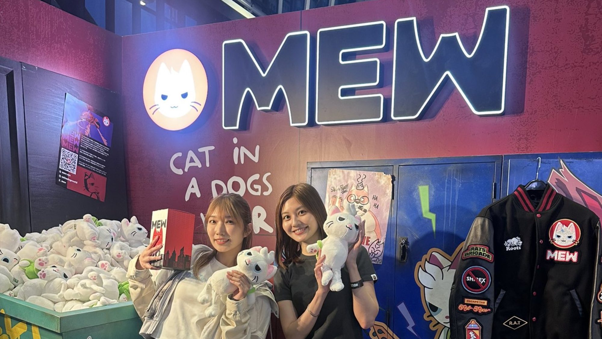 Two fans holding Mew plush toys at a Mew pop-up with a lit MEW sign, a bin of plushies and a Mew varsity jacket